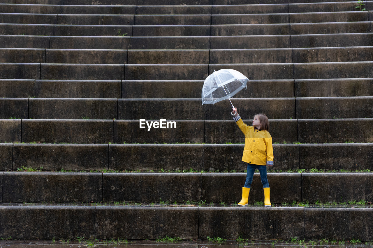 Kid in yellow raincoat and rubber boots playing under umbrella along stone steps on rainy day in city