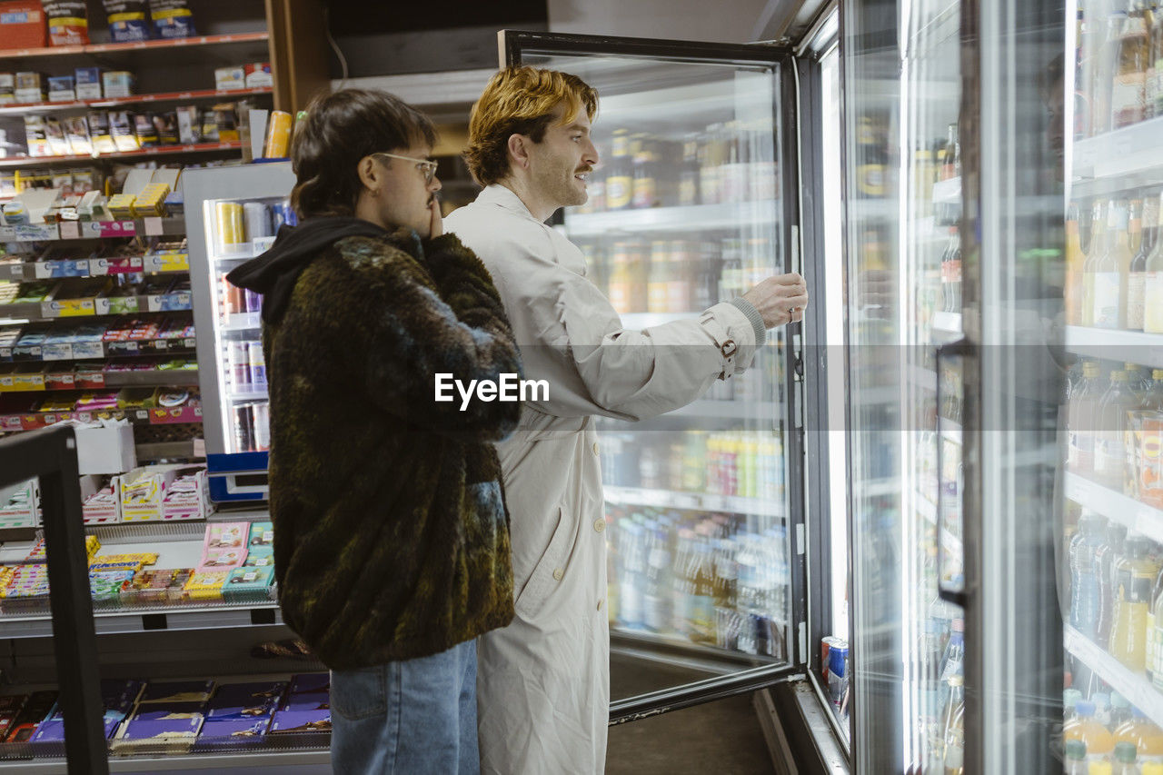 Young gay couple choosing beverages from refrigerated section while doing shopping at supermarket