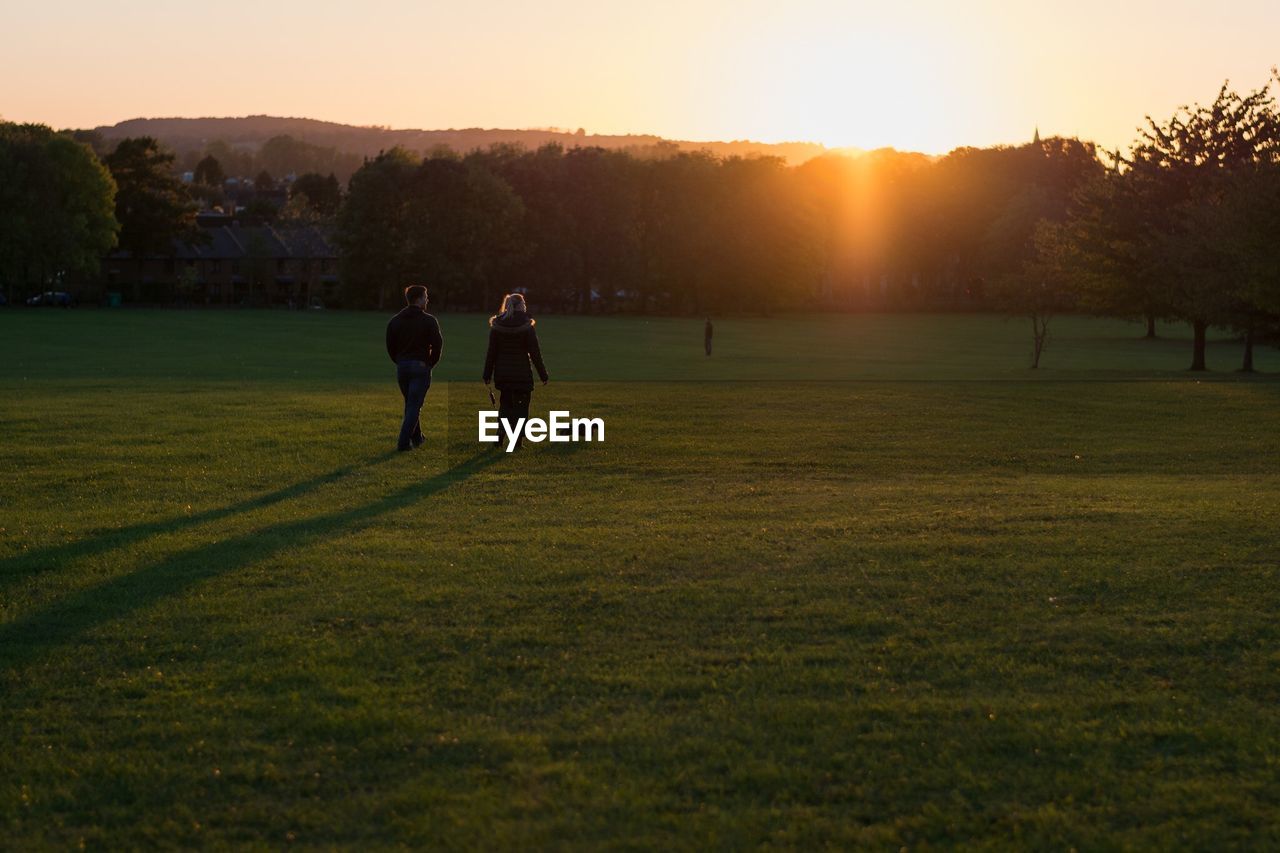 Rear view of man and woman walking with dog in park during sunset