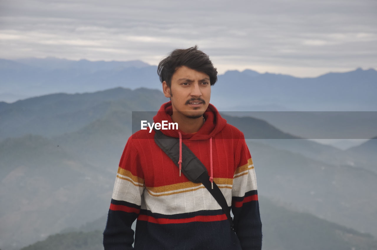 Waist-up of a indian young guy looking sideways while standing against the background of mountains 