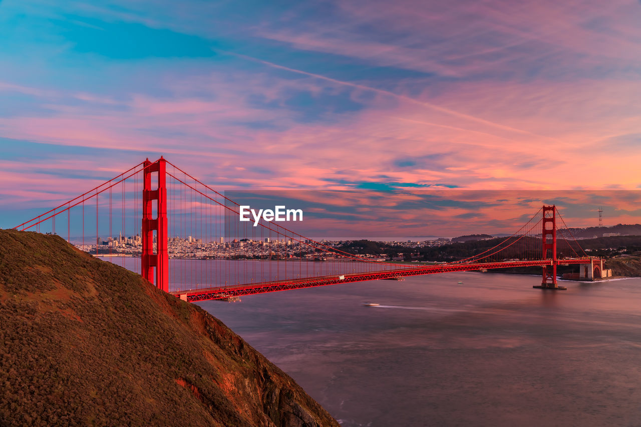 suspension bridge over river against sky at sunset