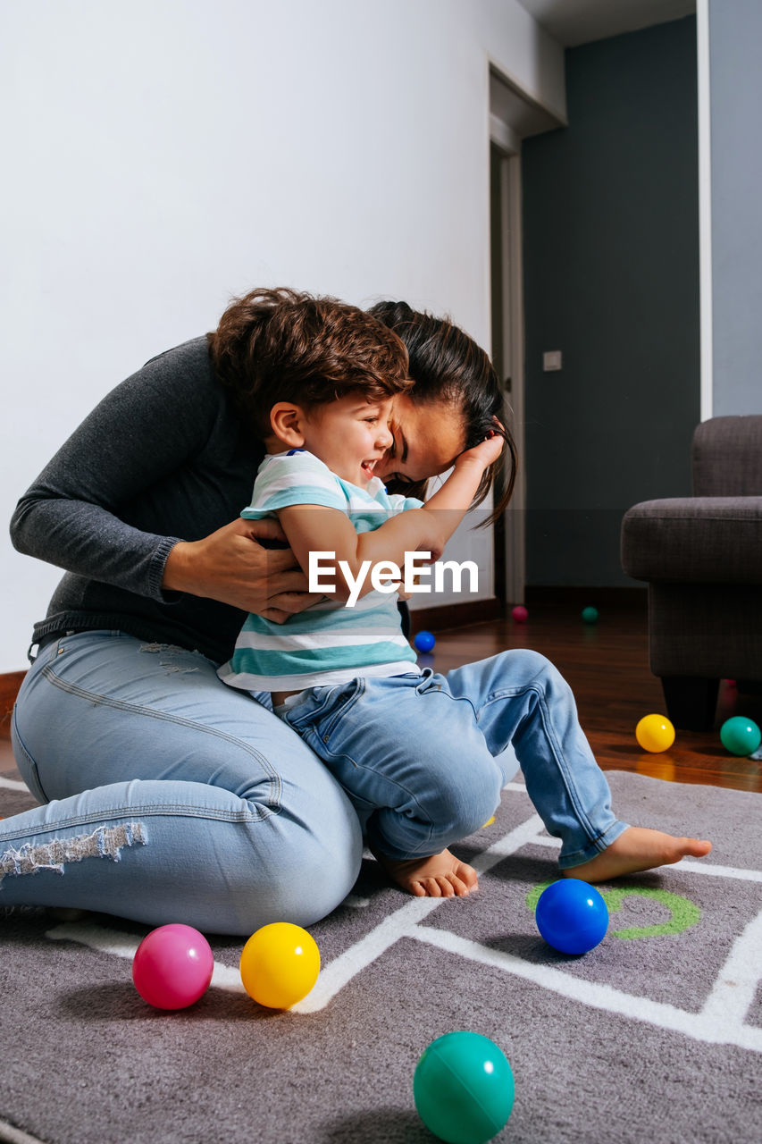 Adult woman tickling and kissing cheerful cute boy while sitting on floor amidst colorful balls and having fun in nursery together