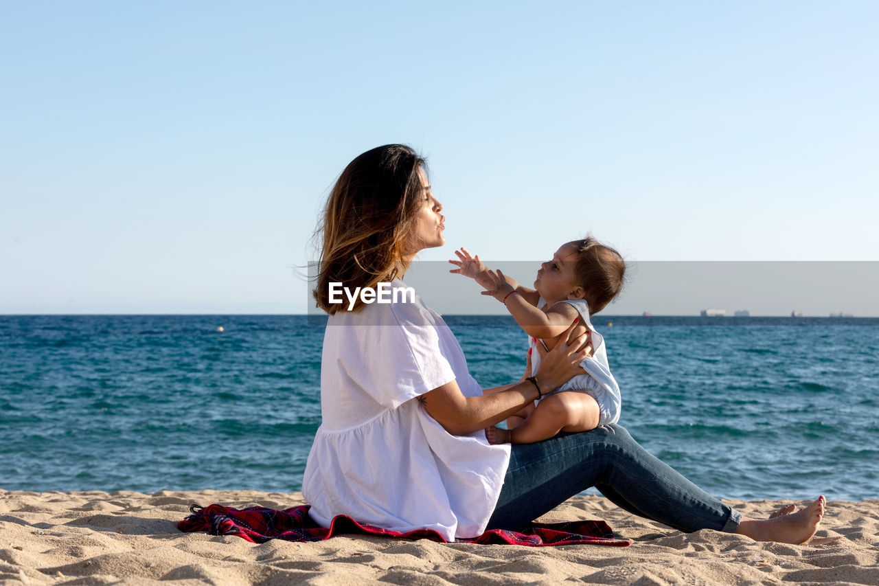 rear view of woman sitting on rock at beach against clear sky