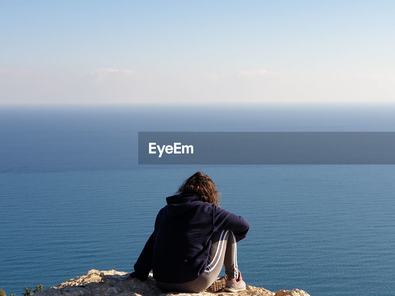 REAR VIEW OF MAN SITTING AT SEA AGAINST SKY