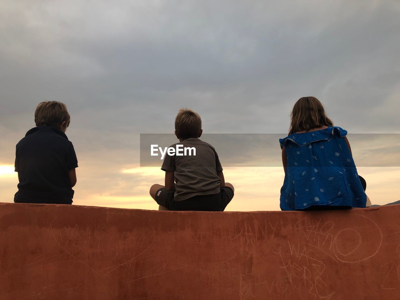 REAR VIEW OF FRIENDS SITTING ON ROCK AGAINST SKY