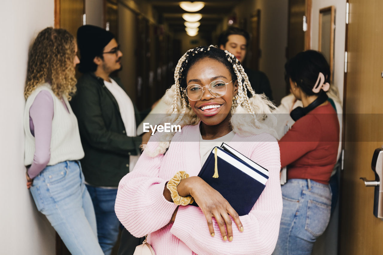 Portrait of smiling young woman with diary while multiracial friends in background at corridor in college dorm