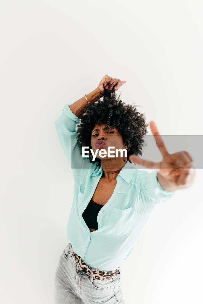 Playful young african american female in trendy outfit having fun and showing peace sign on white background