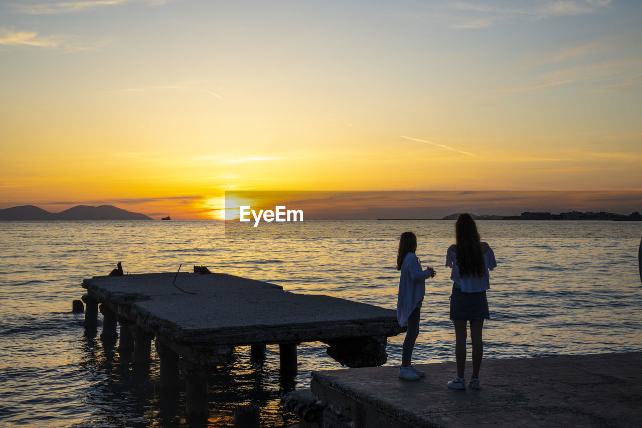 REAR VIEW OF WOMEN STANDING ON BEACH DURING SUNSET