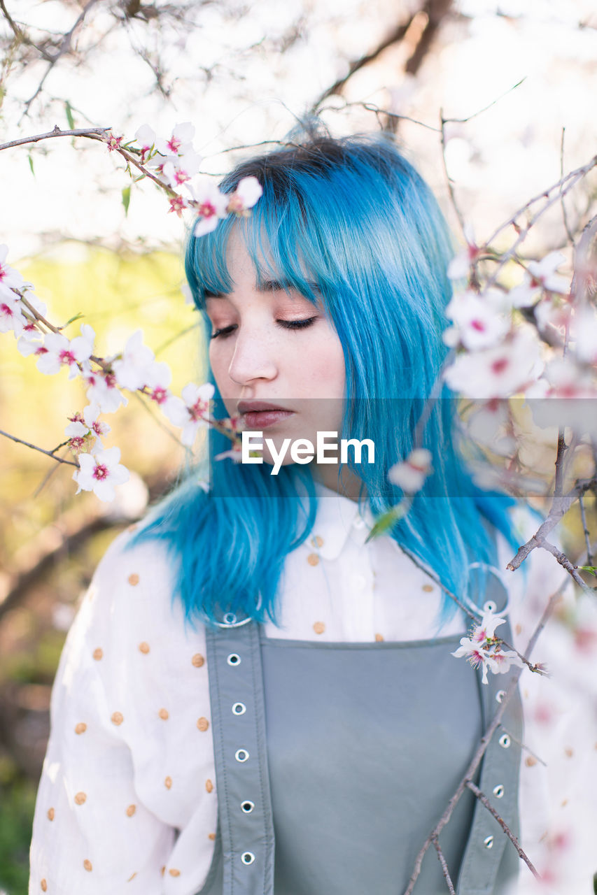 Young stylish female with long blue hair with closed eyes wearing trendy overall enjoying fragrance of blooming tree while standing in spring garden