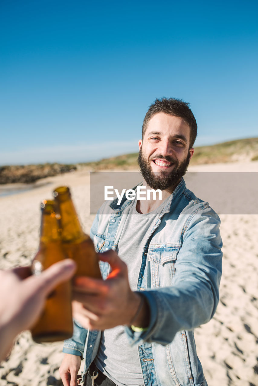 Smiling young man toasting alcoholic drink bottle with friend at beach during sunny day
