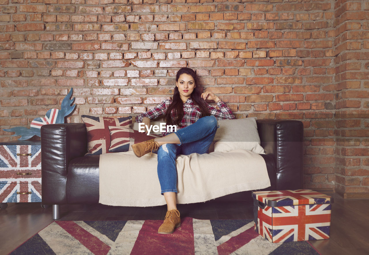 FULL LENGTH PORTRAIT OF YOUNG WOMAN SITTING ON SOFA AGAINST BRICK WALL