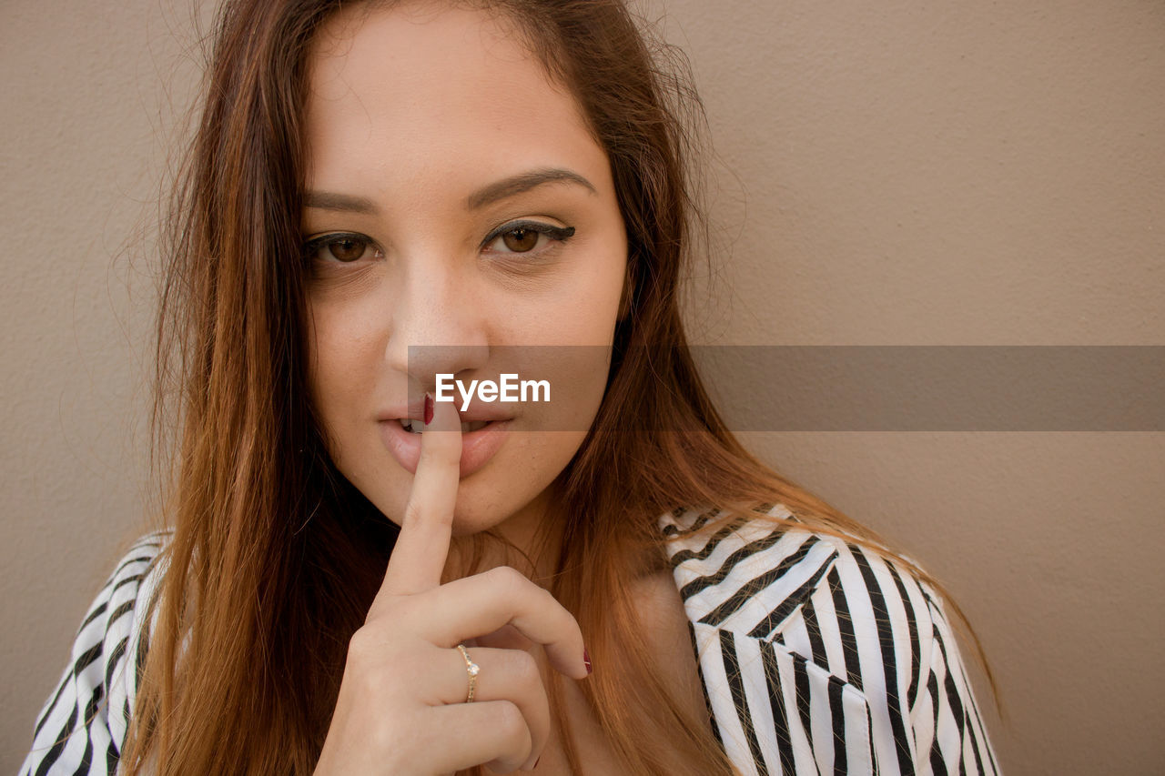 Close-up portrait of beautiful woman with finger on lips standing against brown wall