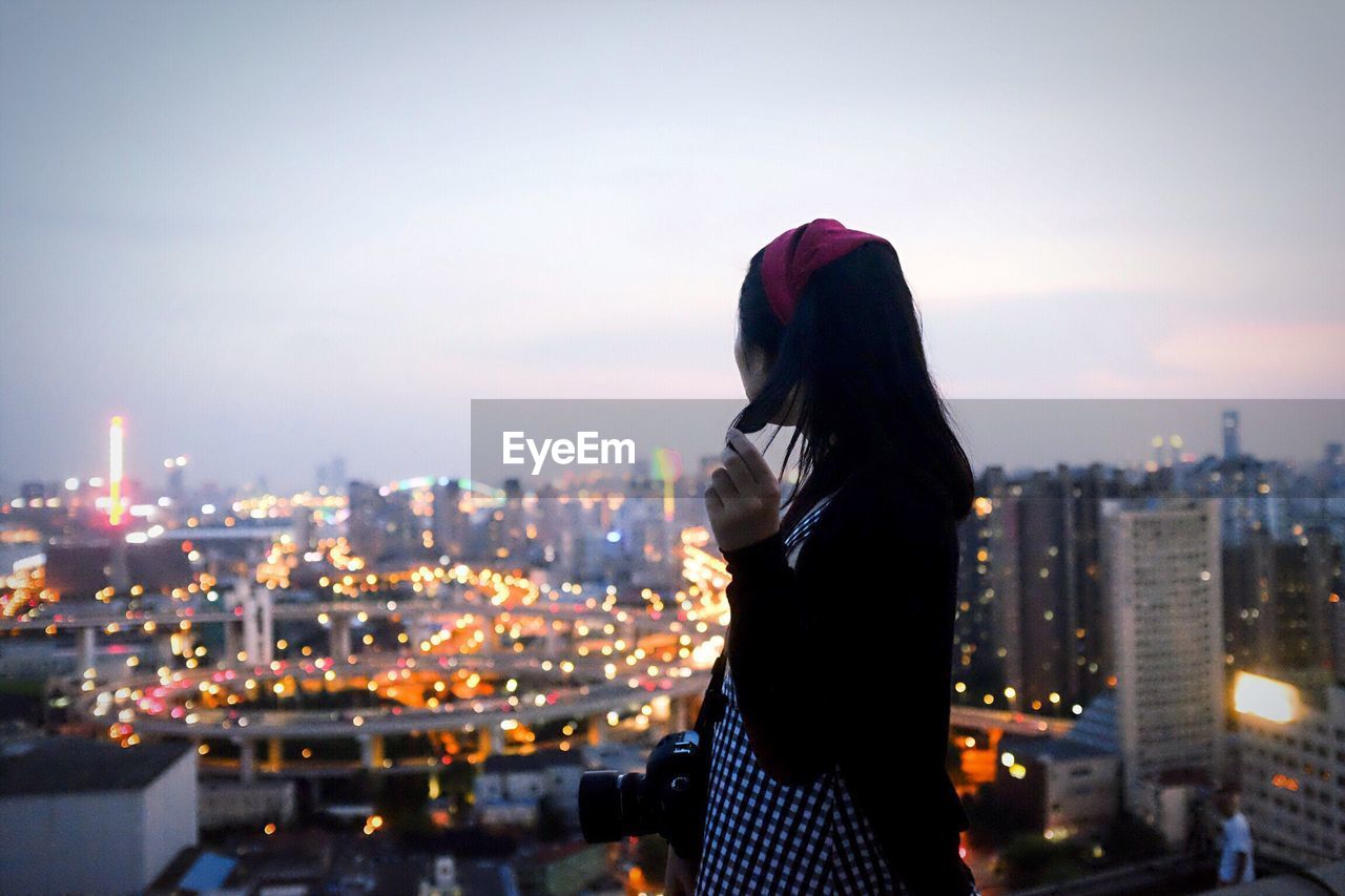 REAR VIEW OF WOMAN STANDING BY ILLUMINATED CITYSCAPE AGAINST SKY