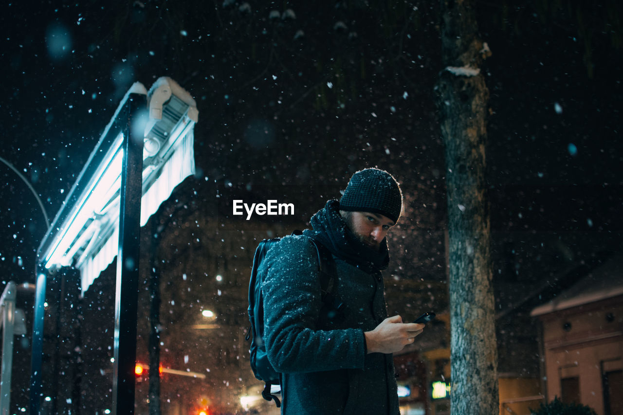 Man using mobile phone while standing against building during snowfall at night