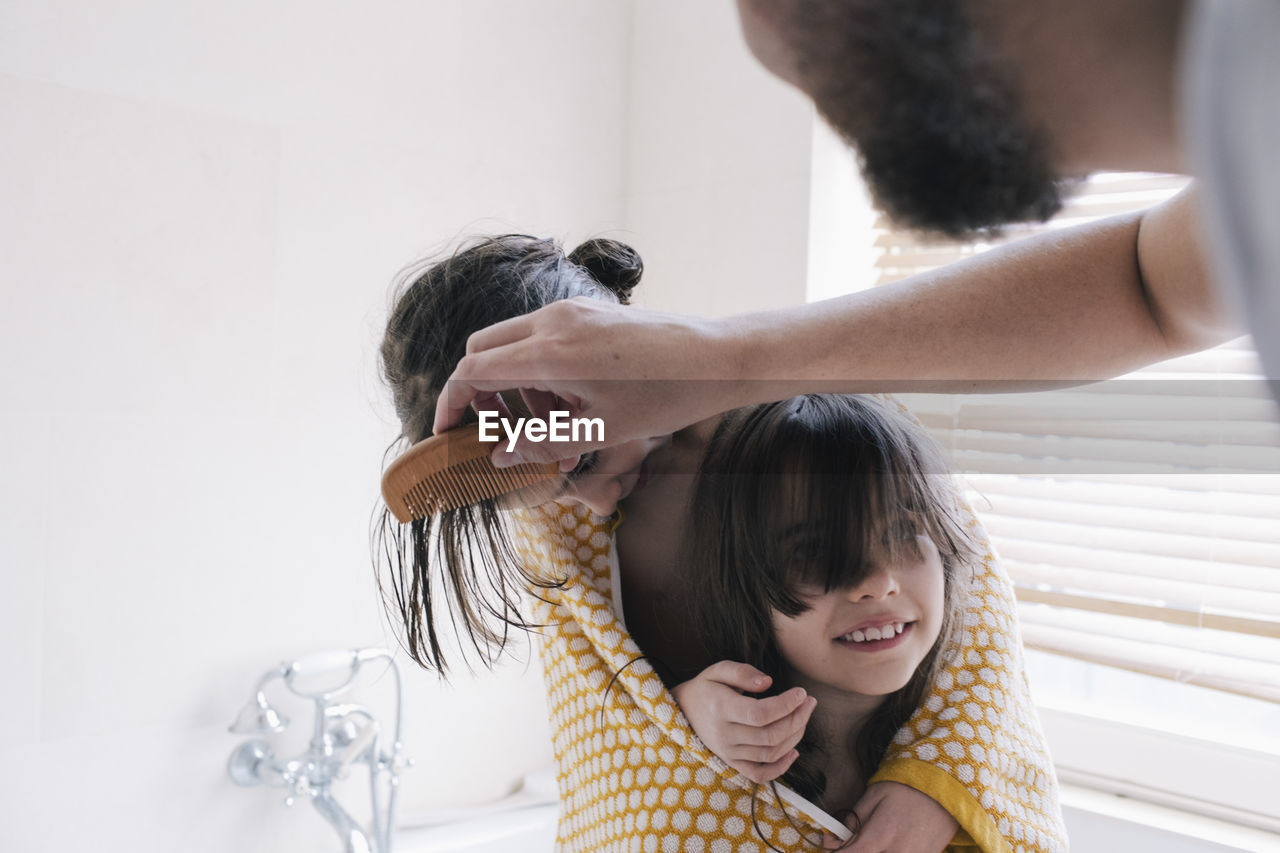 Man combing hair of daughter in towel with sister in bathroom