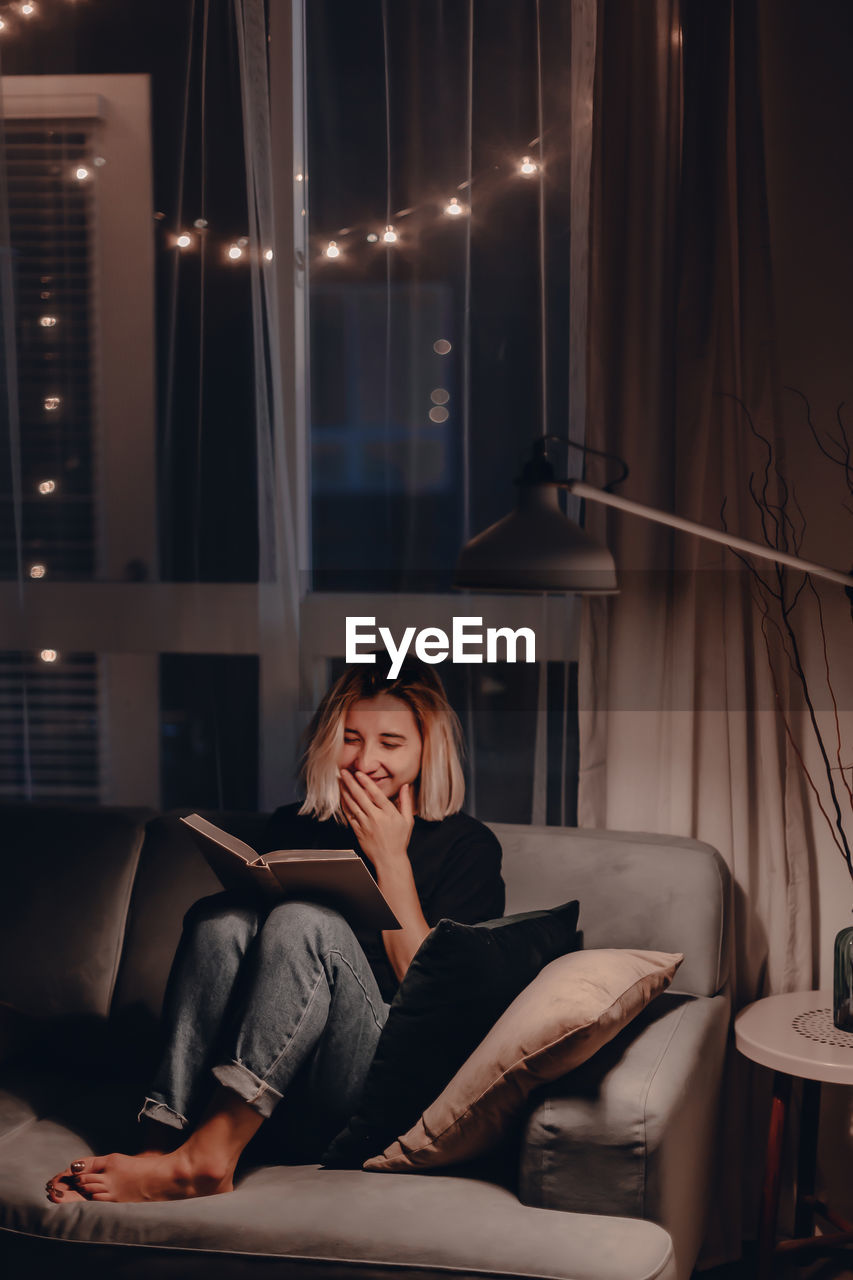 Smiling woman reading book while sitting on sofa at home