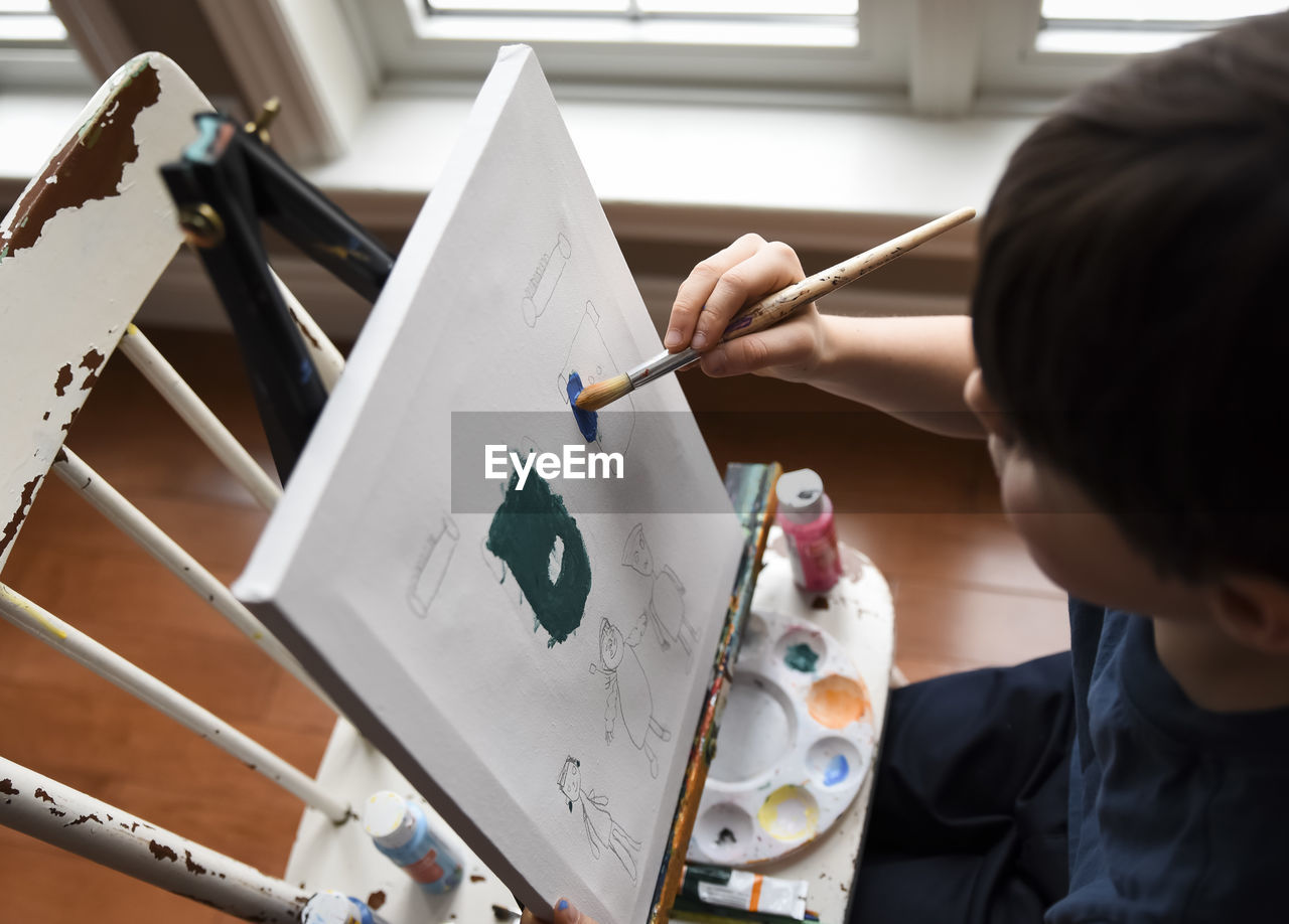 High angle close up of young boy painting on a canvas on an easel.