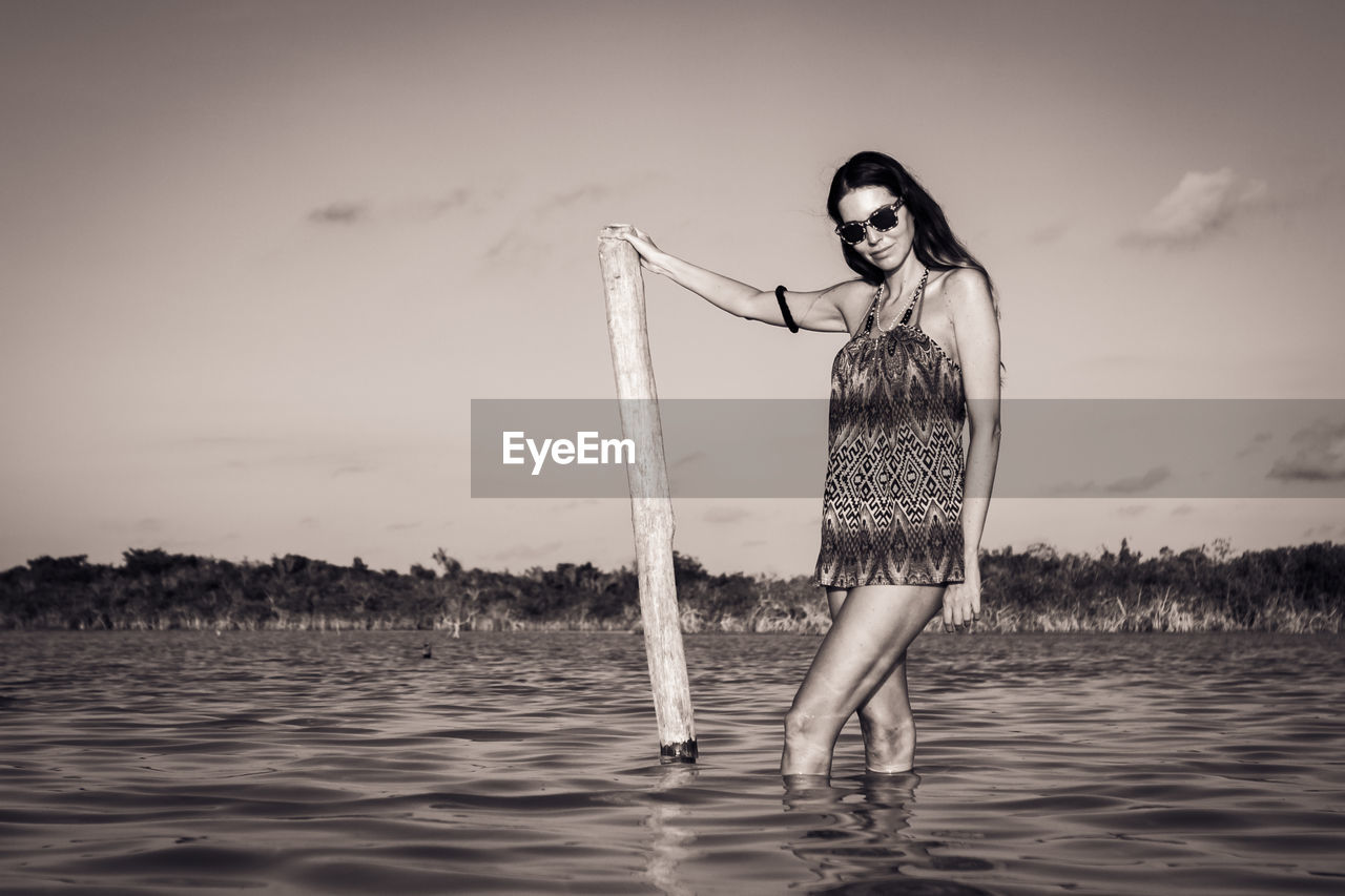 Portrait of young woman wearing sunglasses while standing in sea against sky