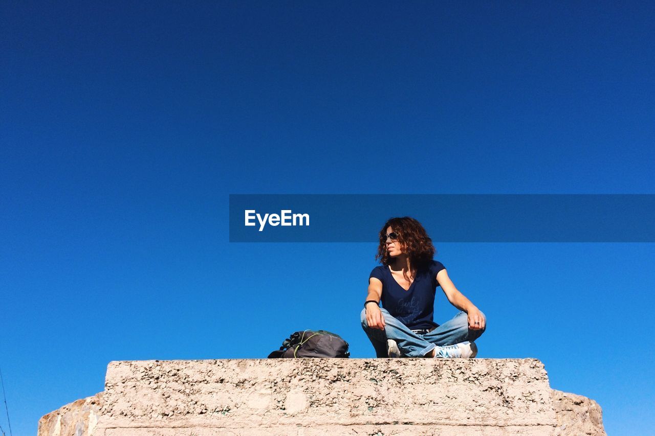 Low angle view of woman sitting on rock against clear blue sky