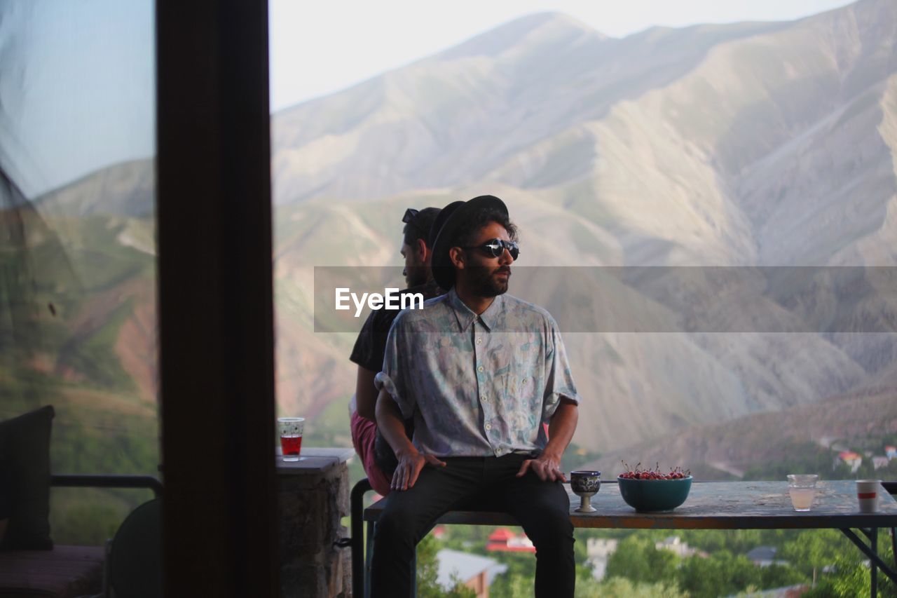 Male friends sitting back to back on bench against mountains