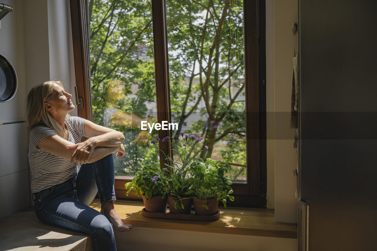 Mature woman enjoying sunlight sitting by window in kitchen