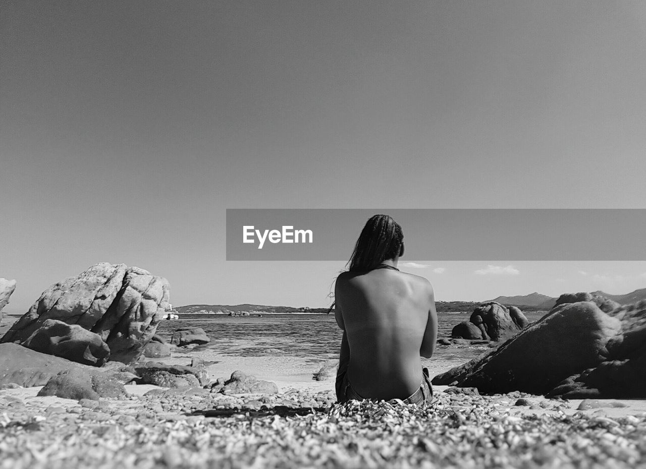 Rear view of topless sensuous woman sitting at beach against sky