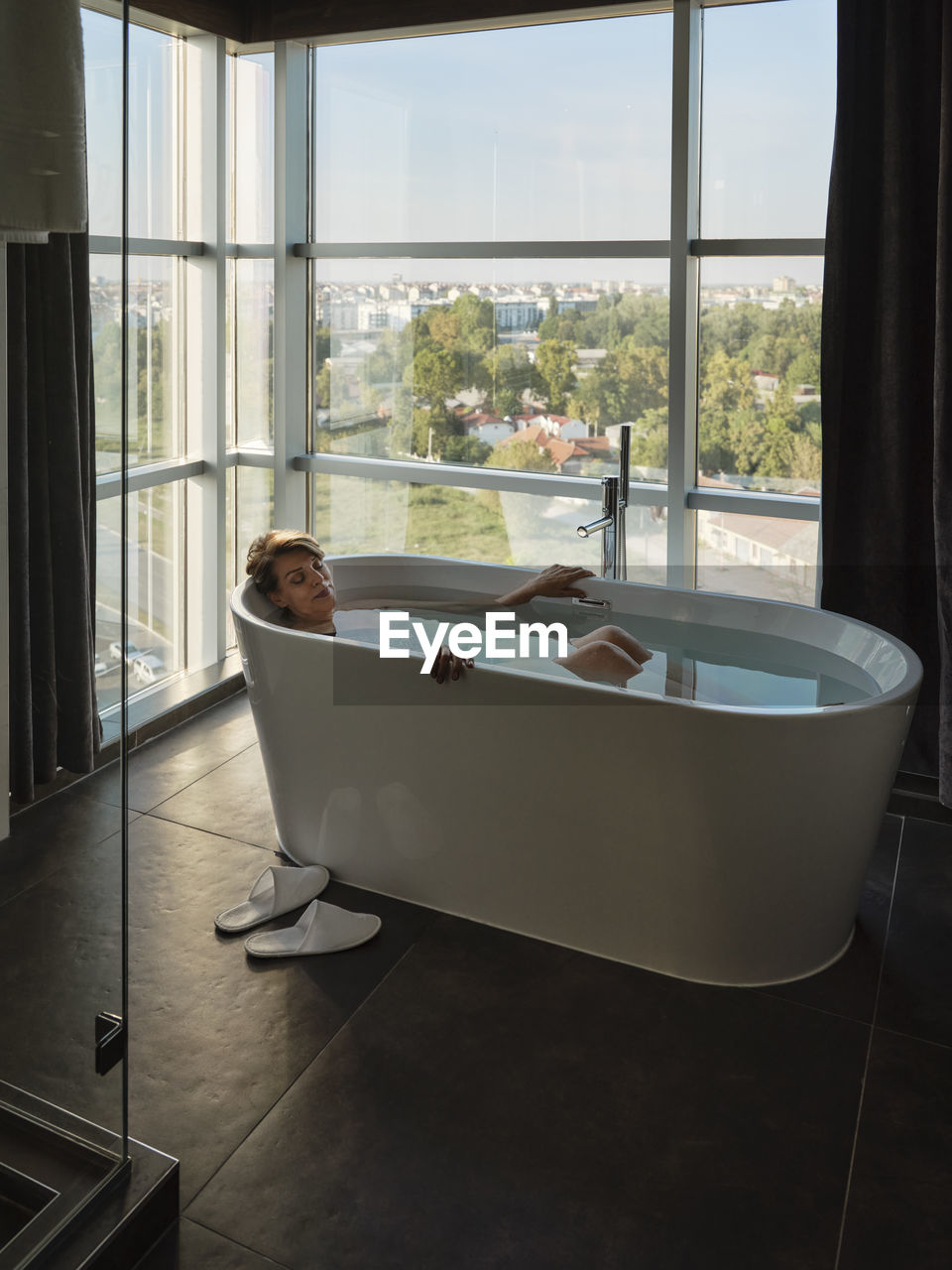 Woman taking bath in bathtub against window at luxury hotel room