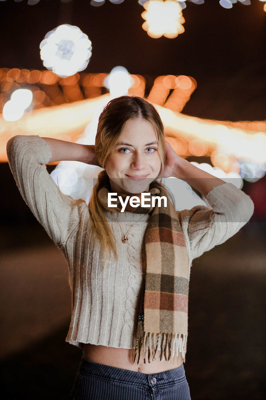 Portrait of young woman standing against illuminated lights at night