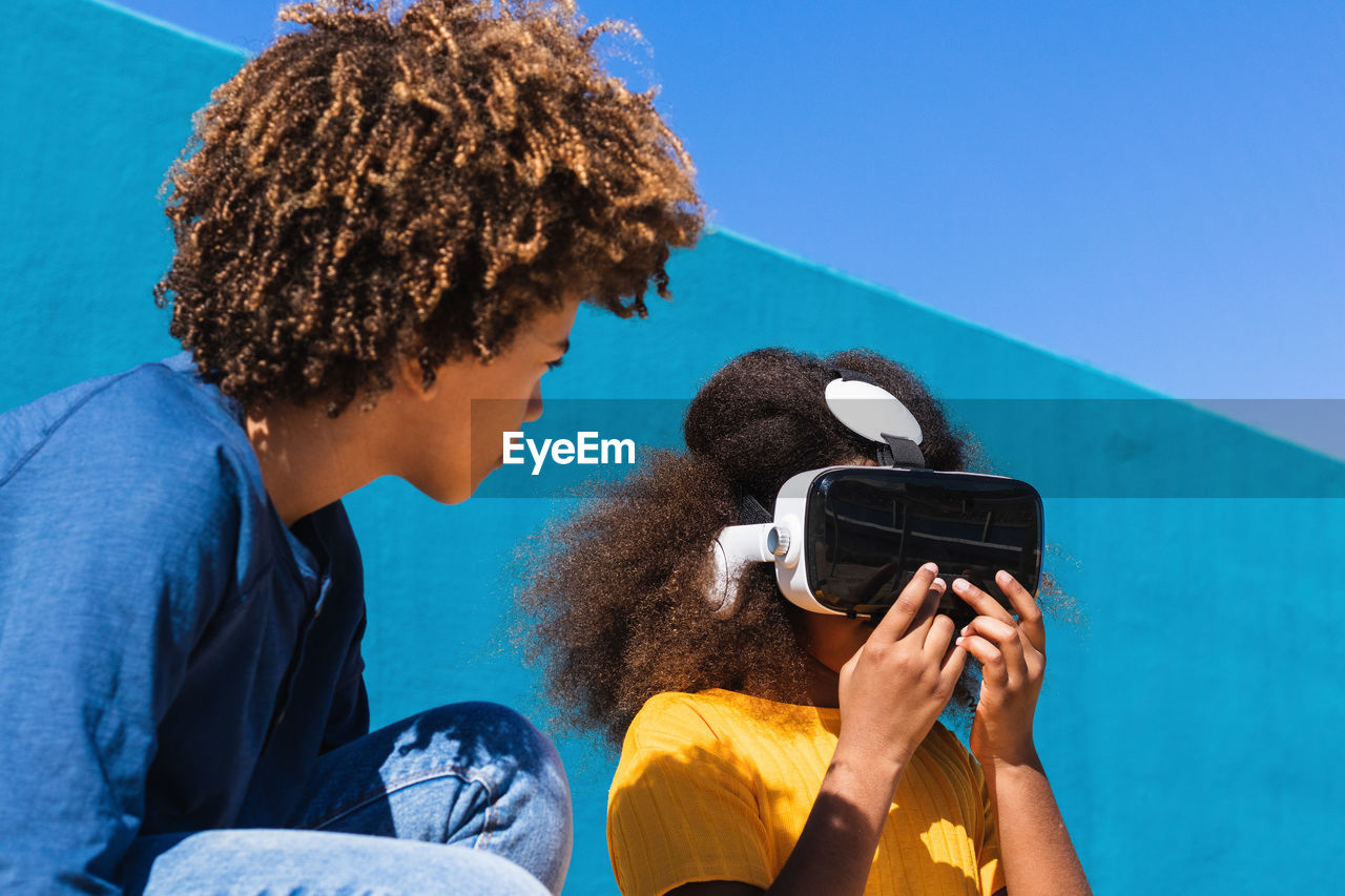 Low angle full body of african american boy communicating with girlfriend wearing vr headset while playing virtual game near colorful wall