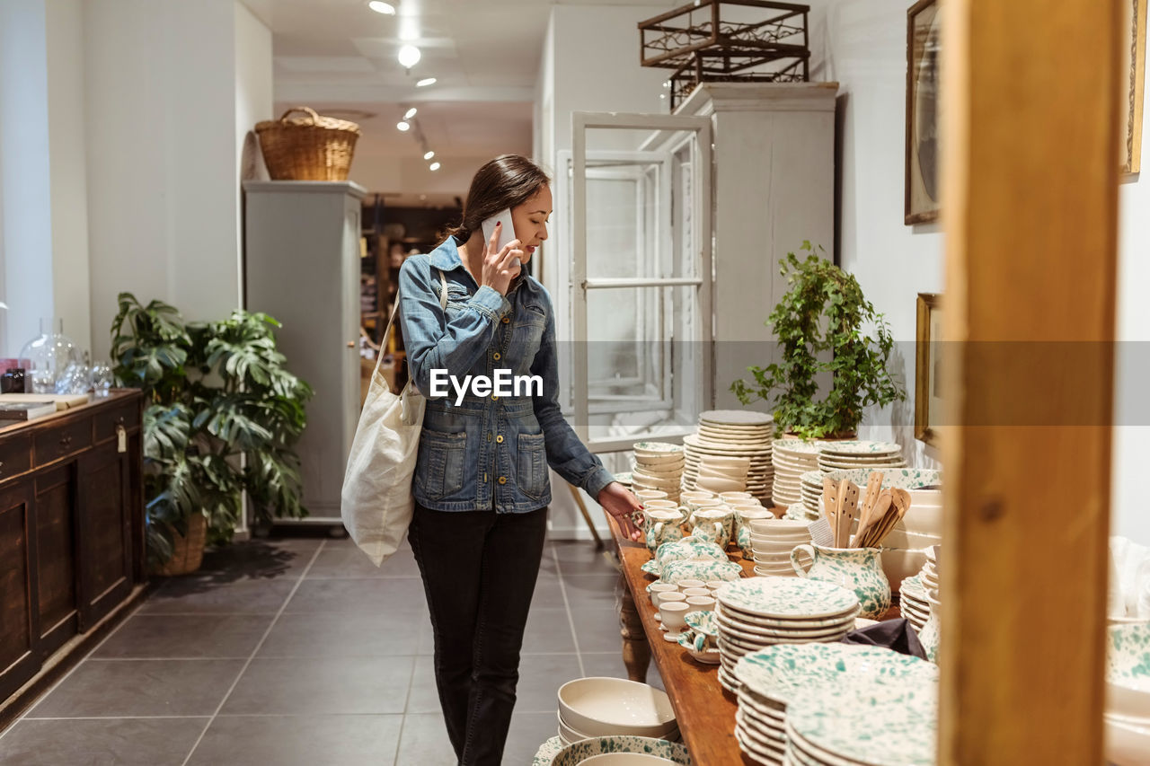 Young woman talking on mobile phone while looking at crockery arranged on table in store