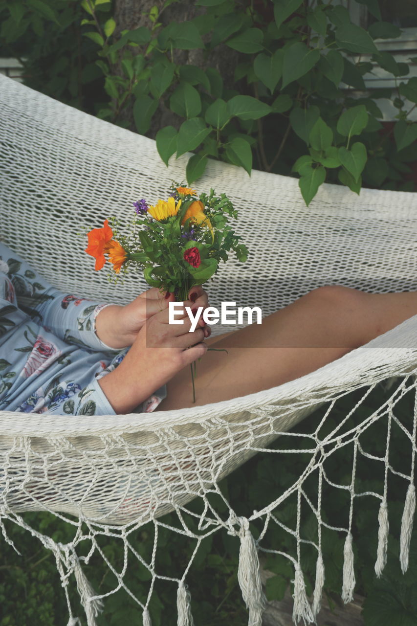 Midsection of woman holding flower bouquet while relaxing in hammock