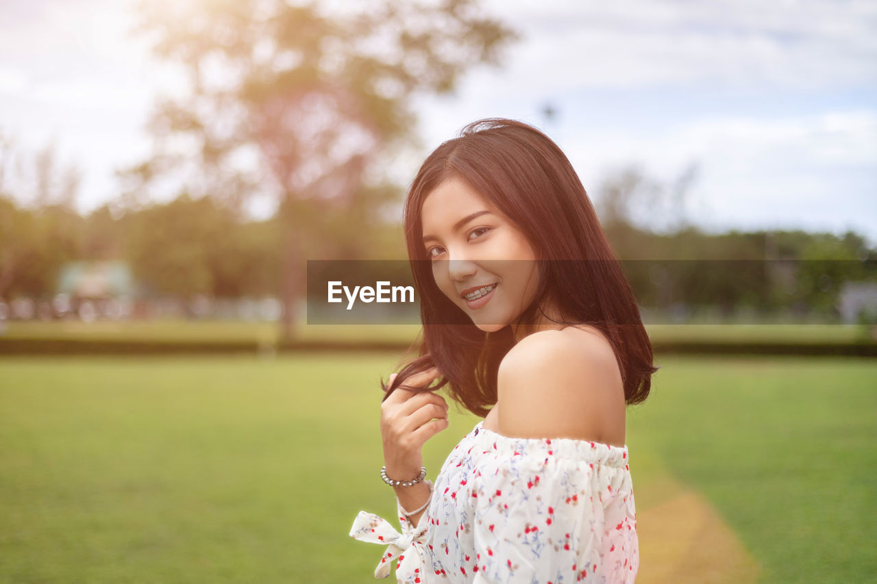 Portrait of smiling young woman standing on grass against trees and sky