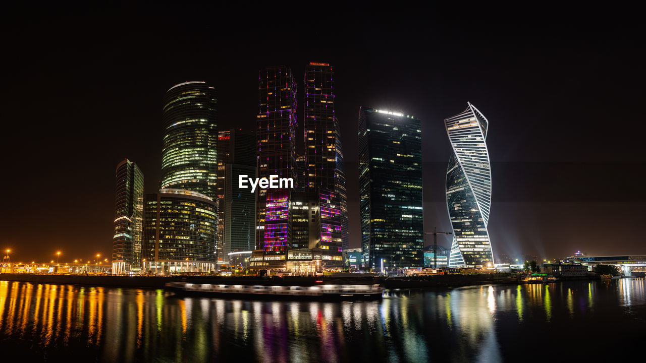 Illuminated modern buildings by river against sky at night