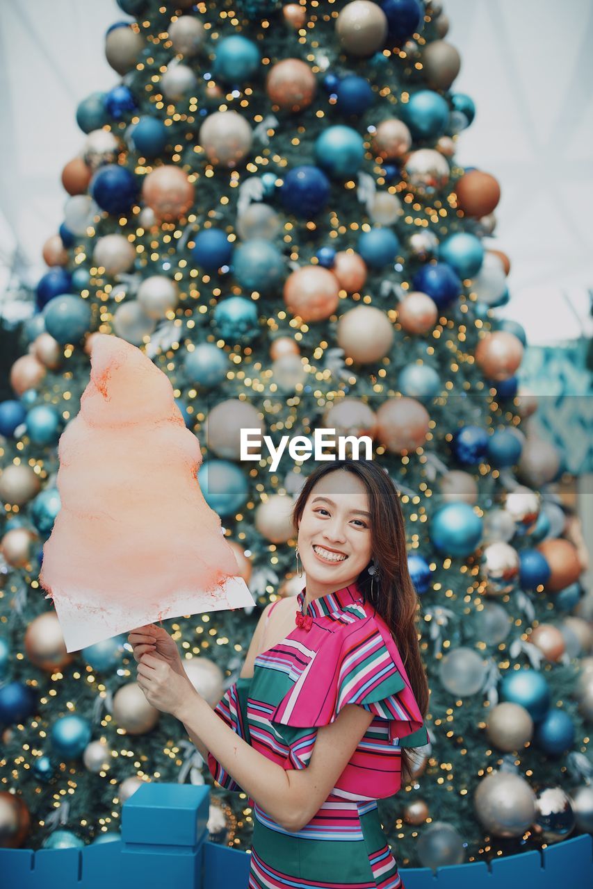 Portrait of smiling young woman standing by christmas tree holding cotton candy outdoors