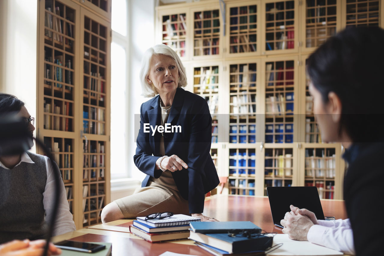 Senior professional gesturing while sitting on table during meeting in library