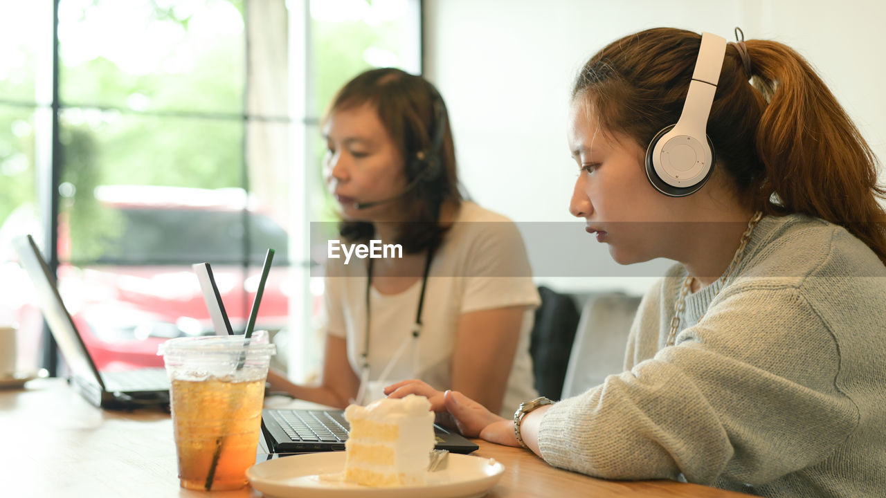 Two young women wearing headphones working with laptops in a cafe, cake and juice on the table.