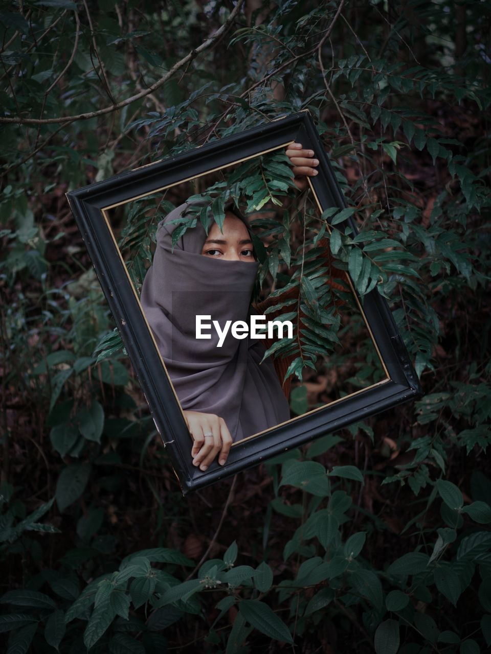 Portrait of woman wearing hijab holding picture frame against plants