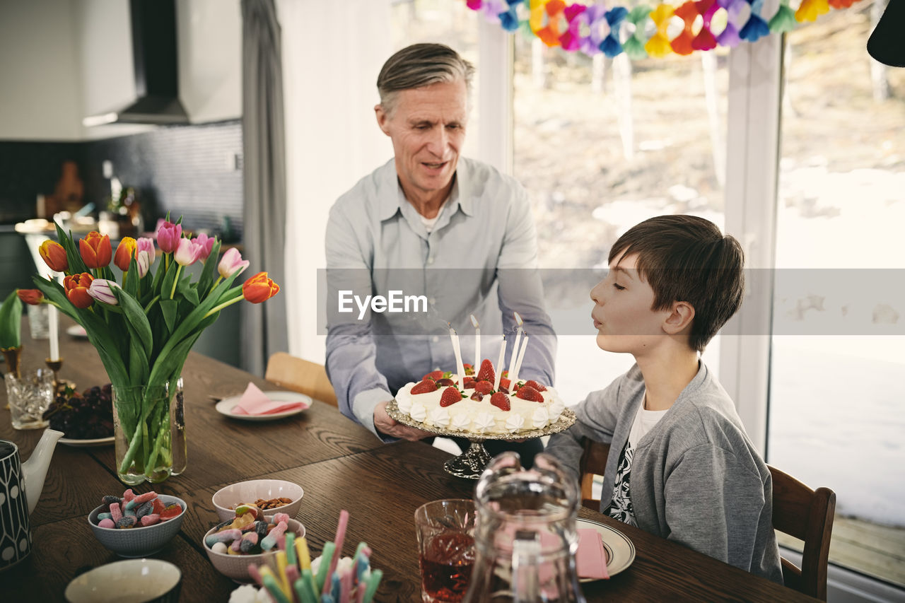 Boy blowing candles while grandfather holding birthday cake at table during party