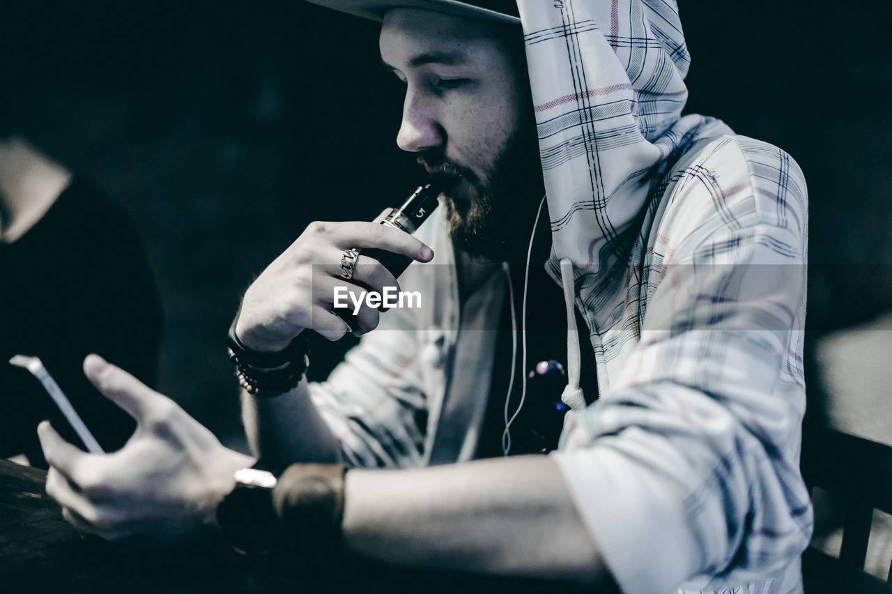 Young man smoking electronic cigarette while using phone at table