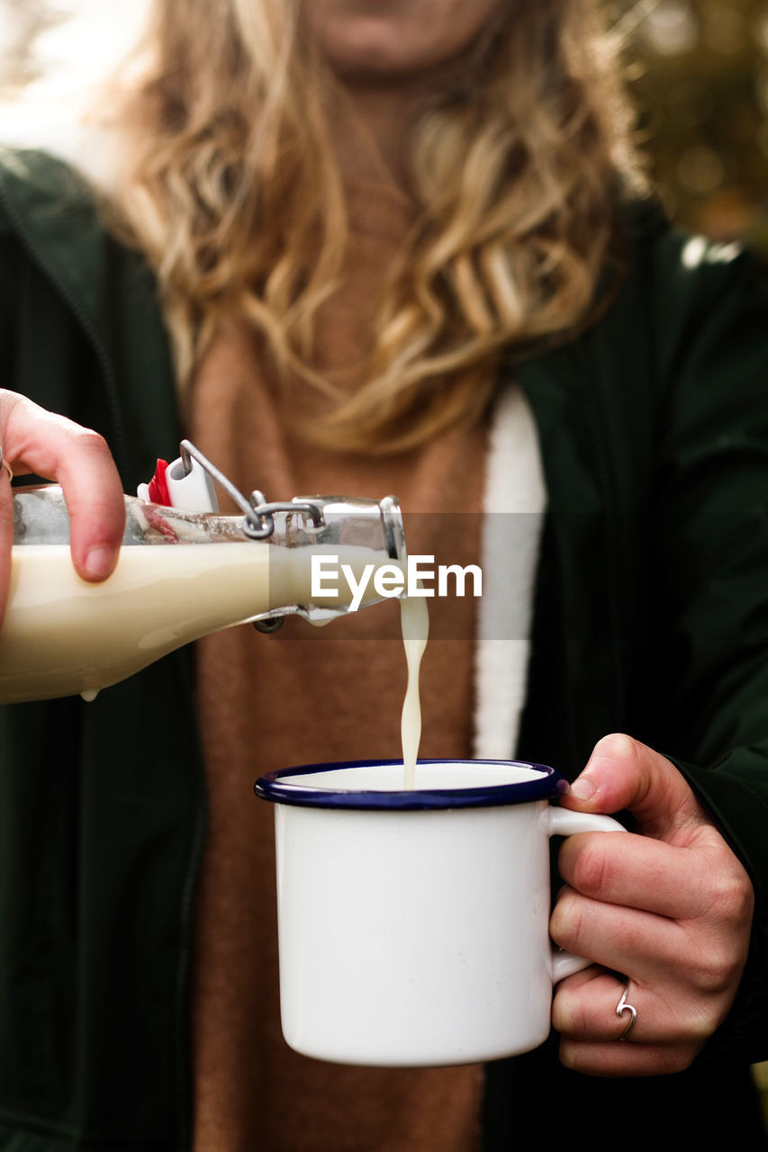 Blond haired faceless woman in casual clothing pouring milk from bottle into mug during picnic