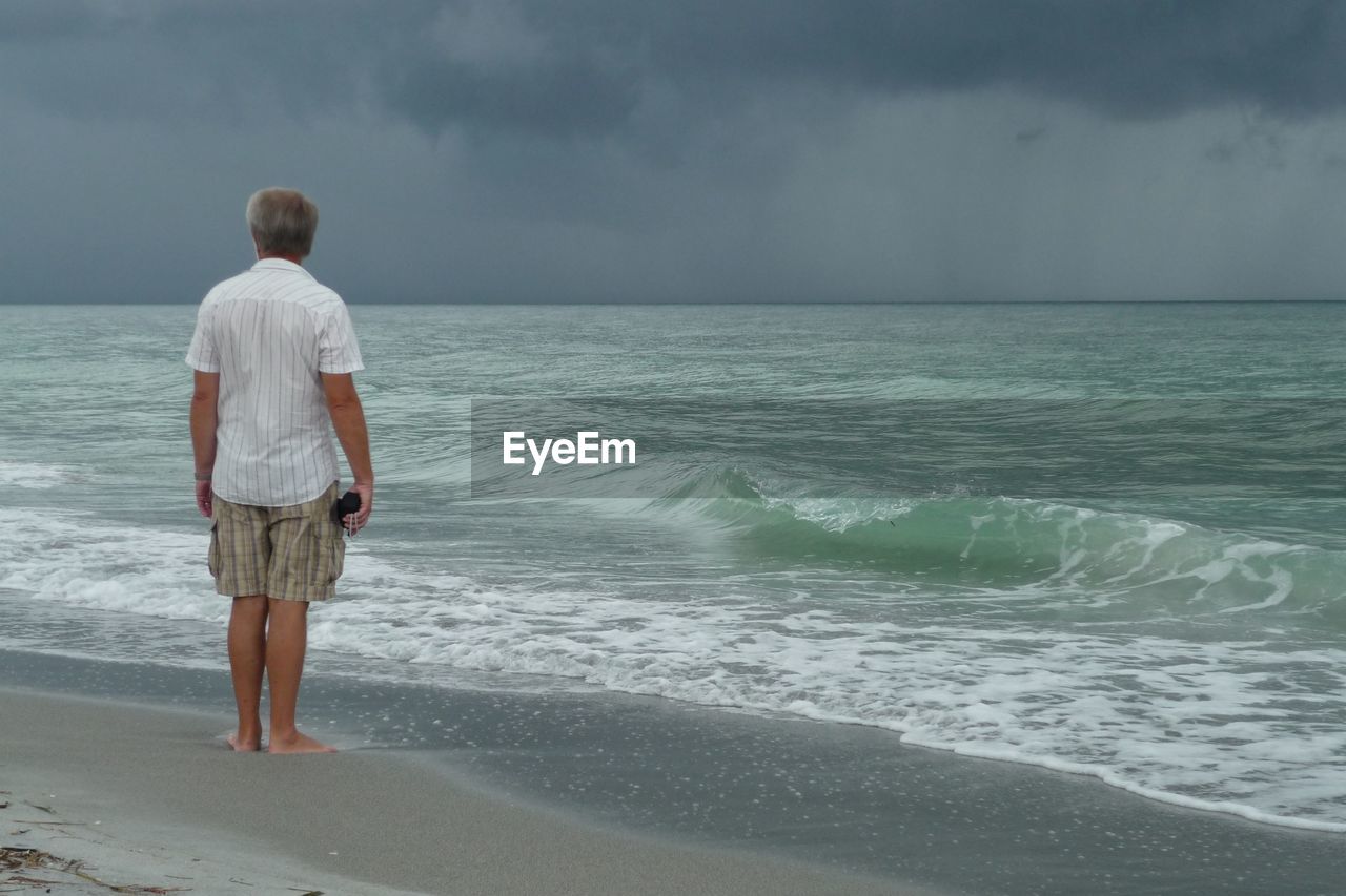 Rear view of man looking at sea against cloudy sky