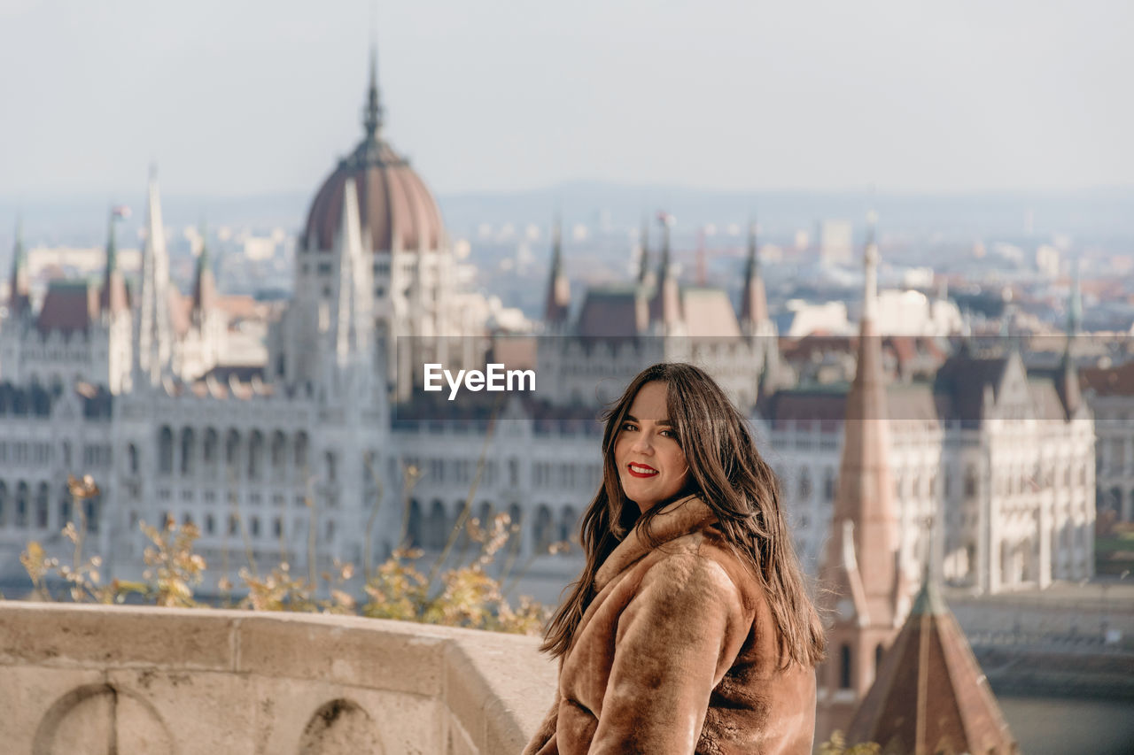 Portrait of beautiful young woman on balcony overlooking hungarian parliament in budapest, hungary