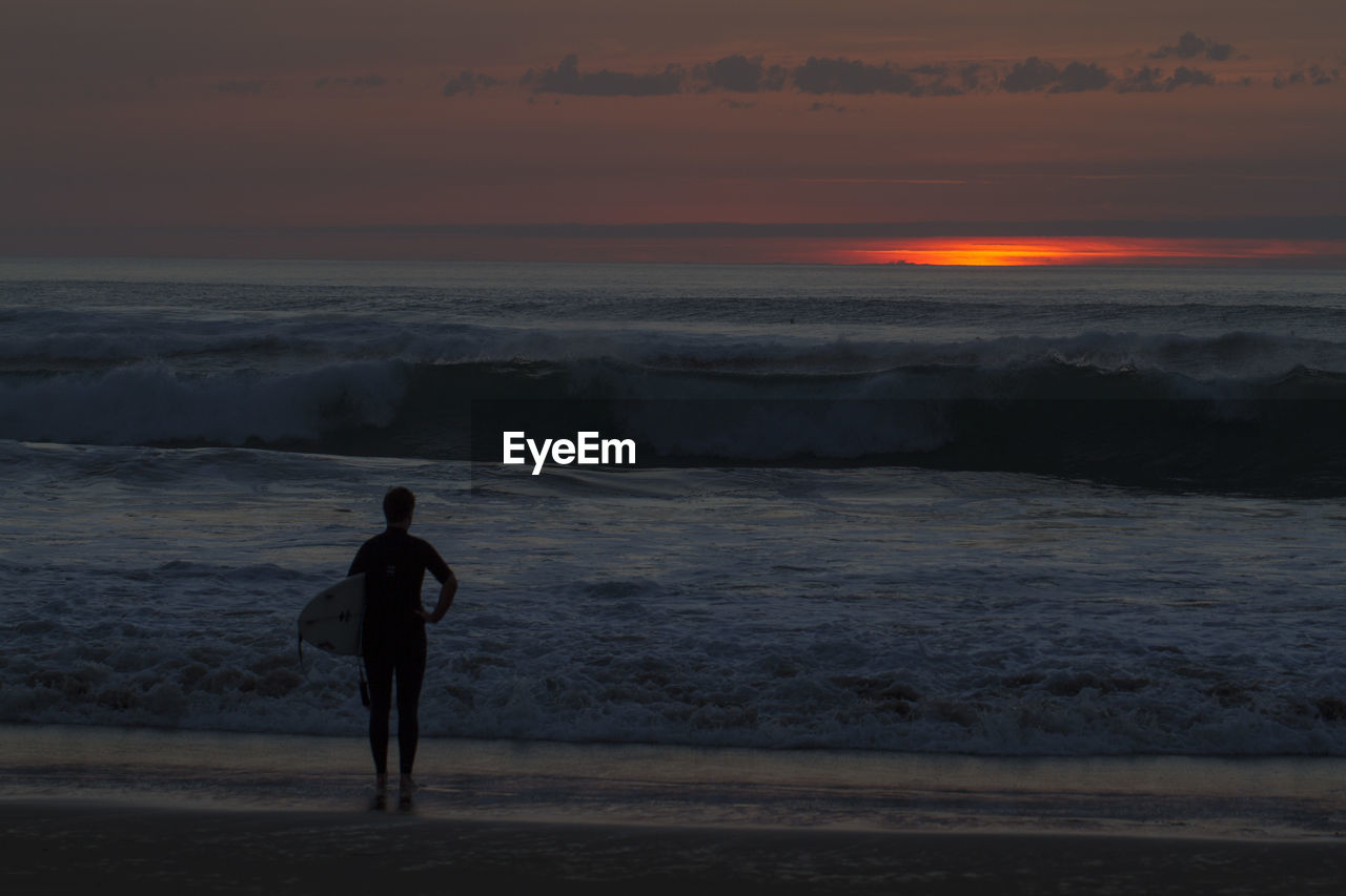 Silhouette man standing with surfboard on shore at beach during sunset