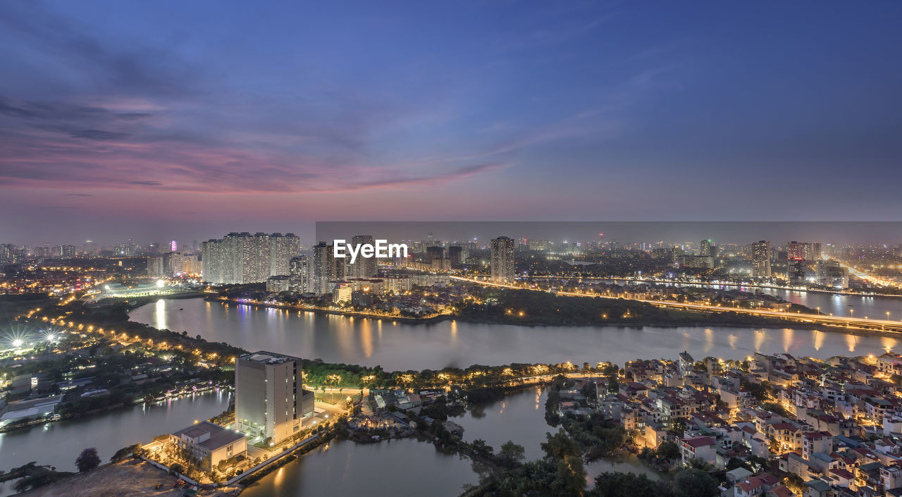 Illuminated city by river against sky at dusk