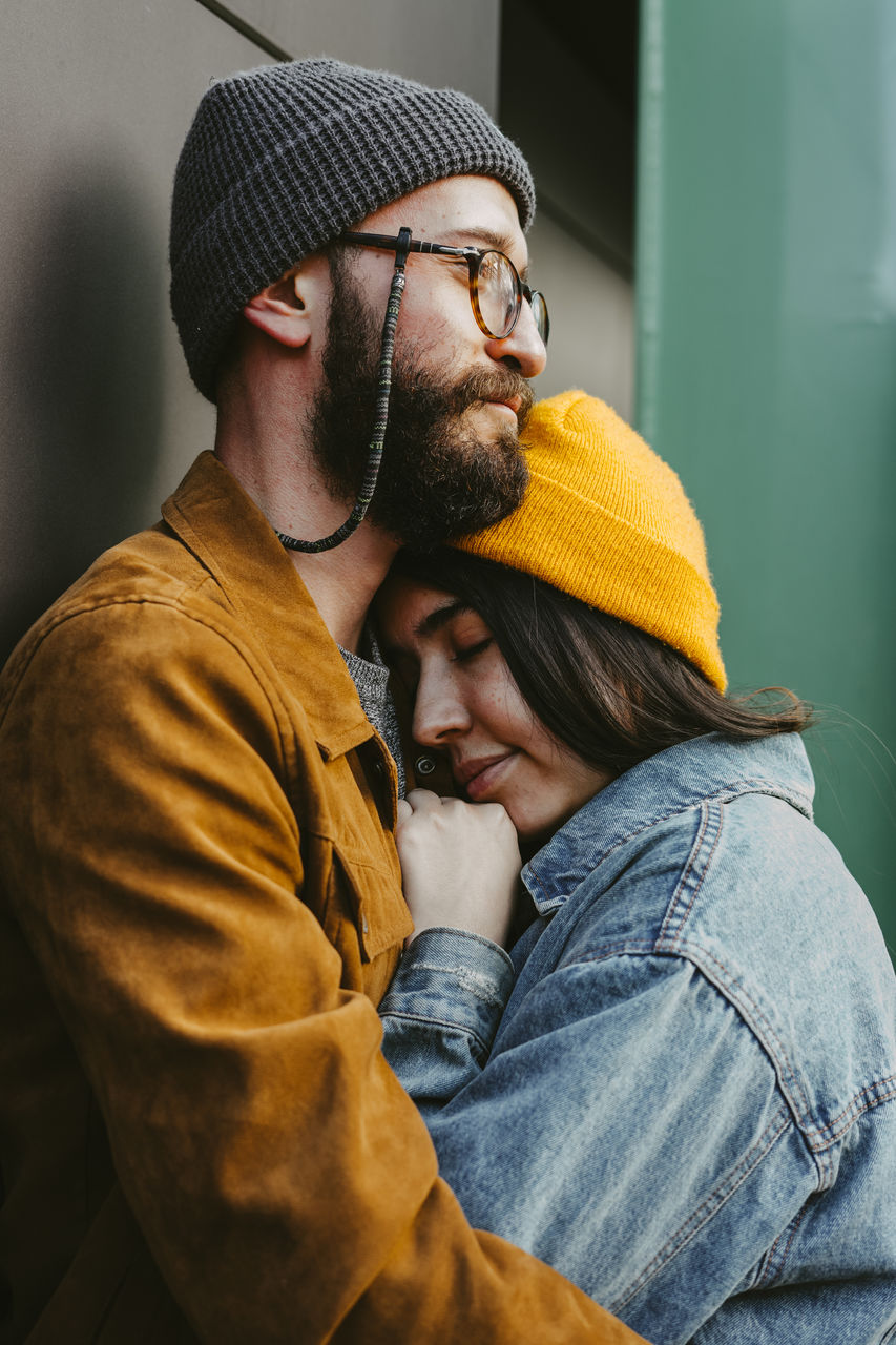 Side view of young stylish couple hugging gently while standing on wooden terrace near house