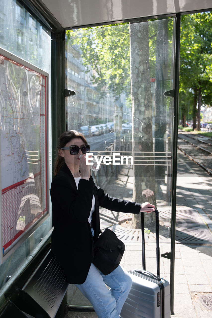 A business woman in sunglasses drinks water at a tram stop in the city, dressed