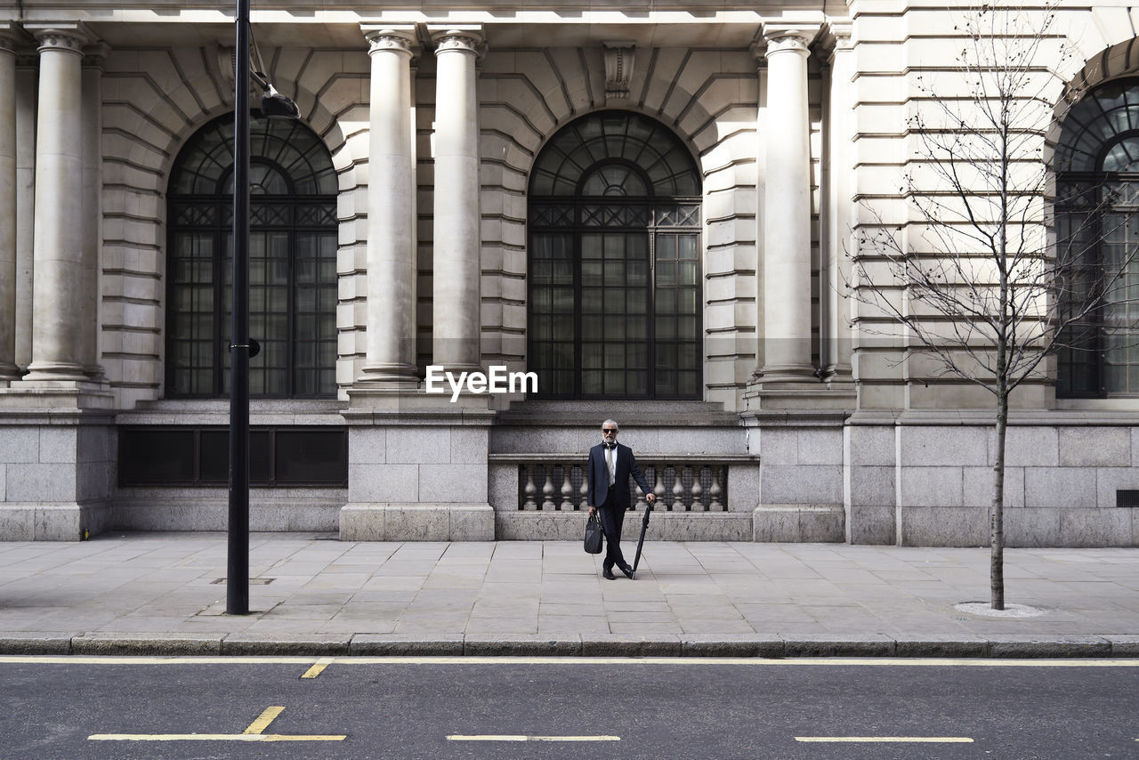 Uk, london, stylish senior businessman with briefcase and umbrella standing on pavement