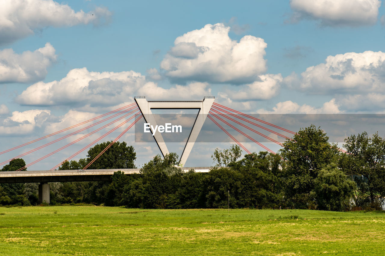 Low angle view of field against sky with a bridge, threes and cloudy sky 