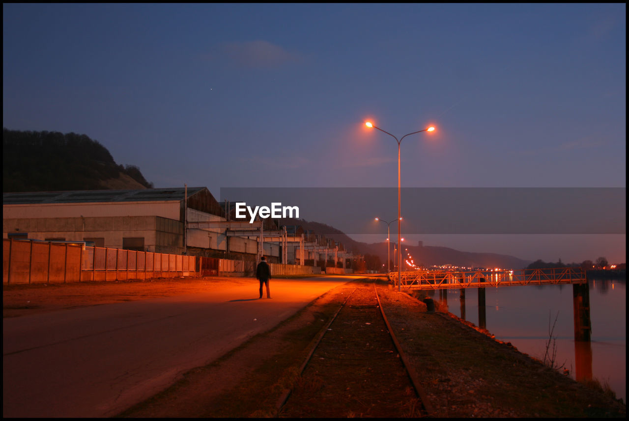 Person walking on road by industry against sky at night