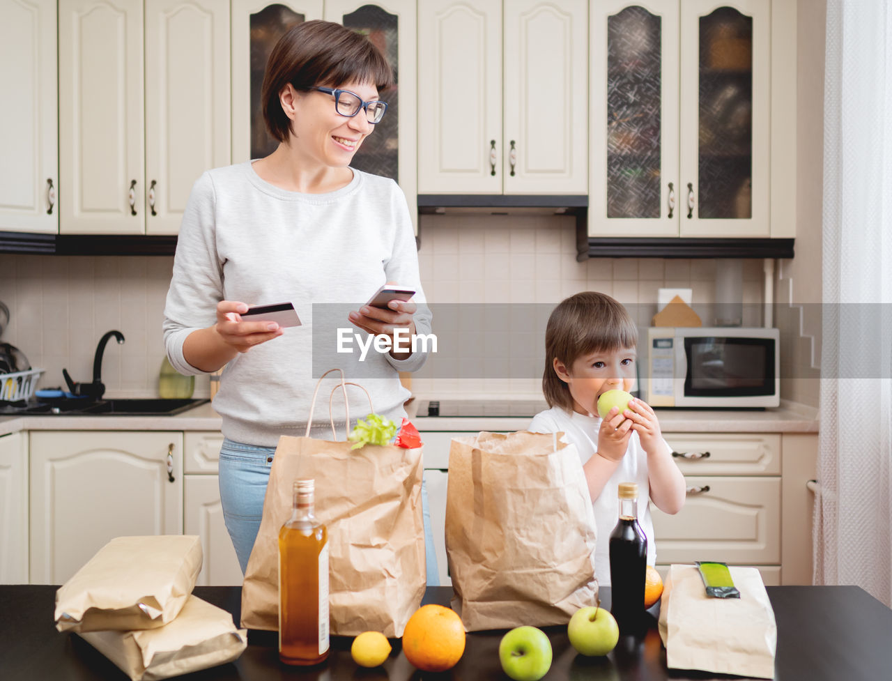 Woman and boy sorts out purchases. kid bites an apple.grocery delivery in paper bags.mother and son.